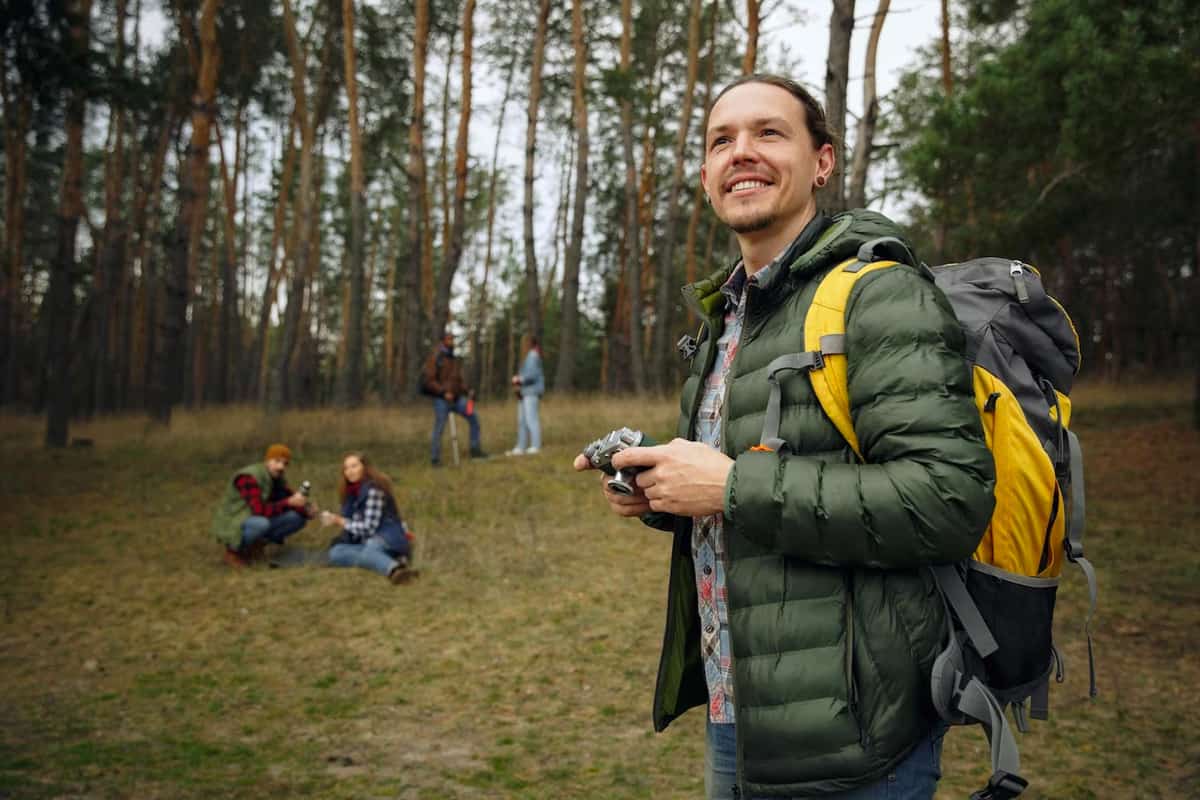 A smiling man with backpack and camera in a forest.