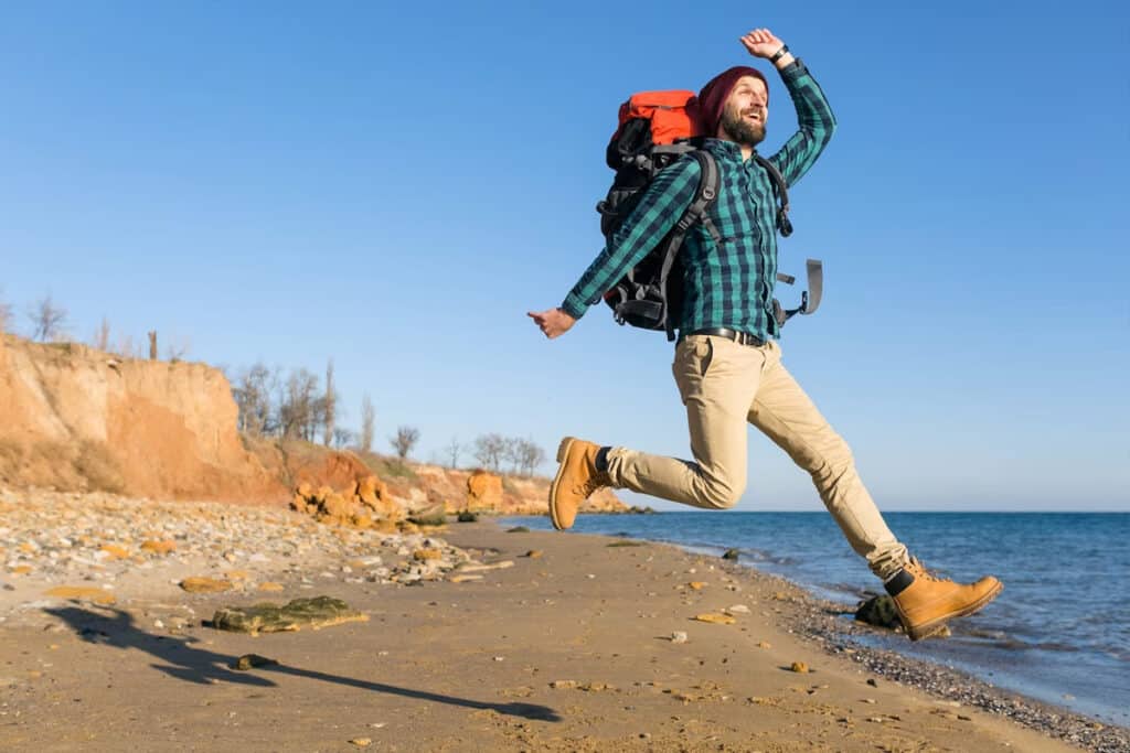 A happy man jumping on the beach with a backpack.